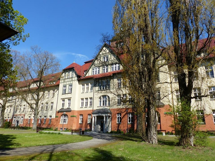 Eingang der Klinik Beelitz, vor dem Gebäude sind grüner Rasen und zahlreiche Bäume.
