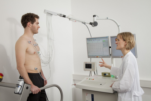 An einem oberkörperfreien Mann sind Elektroden aufgeklebt, eine Ärztin erklärt die Ergebnisse einer Messung, die auf einem Monitor angezeigt werden.