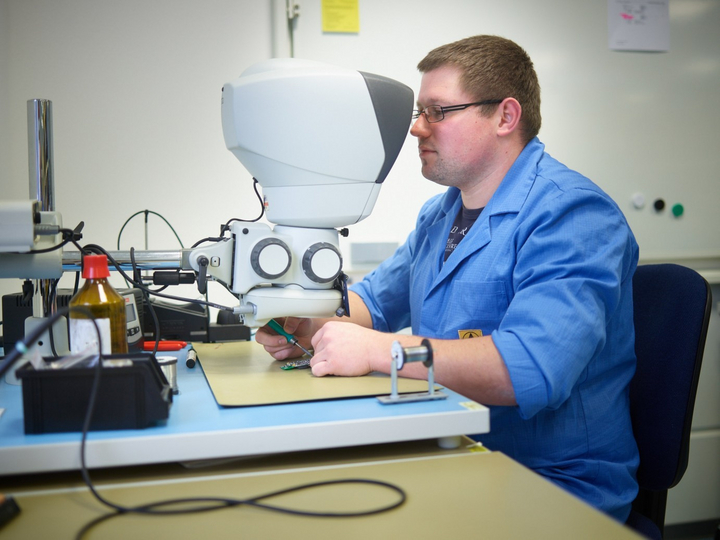 Ein Medizintechniker blickt durch ein Mikroskop und lötet sehr kleine elektronische Bauteile auf eine Platine.