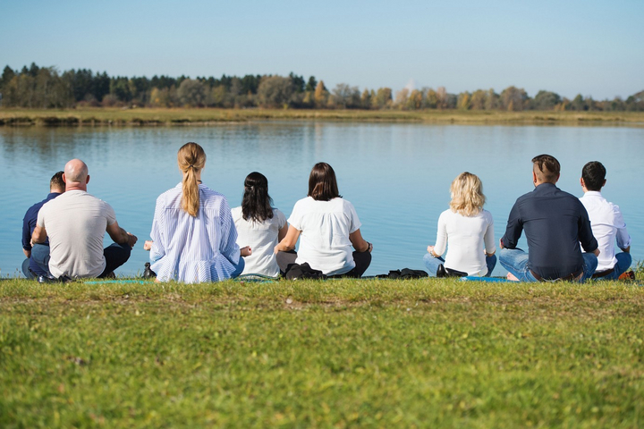 Eine Gruppe von acht Personen meditiert sitzend auf einer Wiese am See.