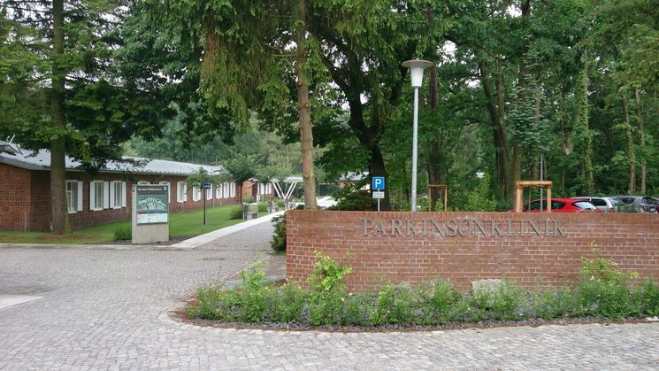 Aufnahme der mit Bäumen bewachsene Einfahrt zum Klinikgelände. Rechts auf der Mauer der Einfahrt steht in großen Buchstaben Parkinsonklinik.