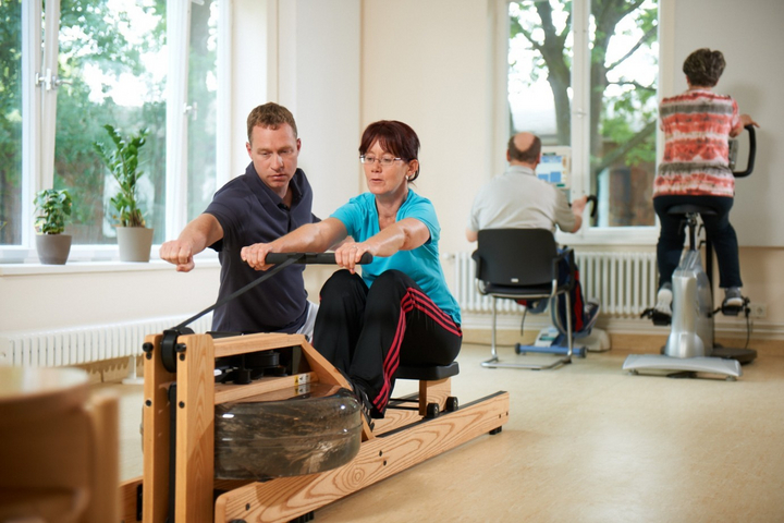 Sporttherapeut trainiert mit einer Patientin am modernen Rudergerät aus Holz. Im Hintergrund trainineren zwei Patienten auf Home Fahrrädern mit Monitoren.