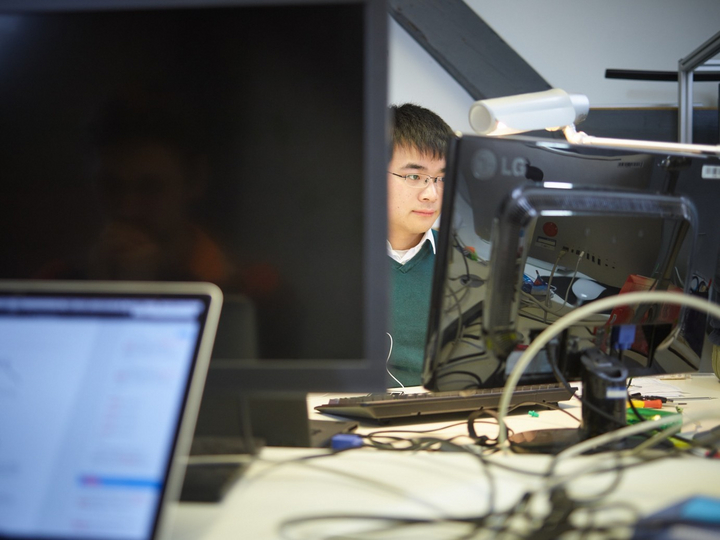 Ein junger Mitarbeitender arbeitet am Bildschirm in einem Büro in dem mehrere Rechner und Laptops zu sehen sind.